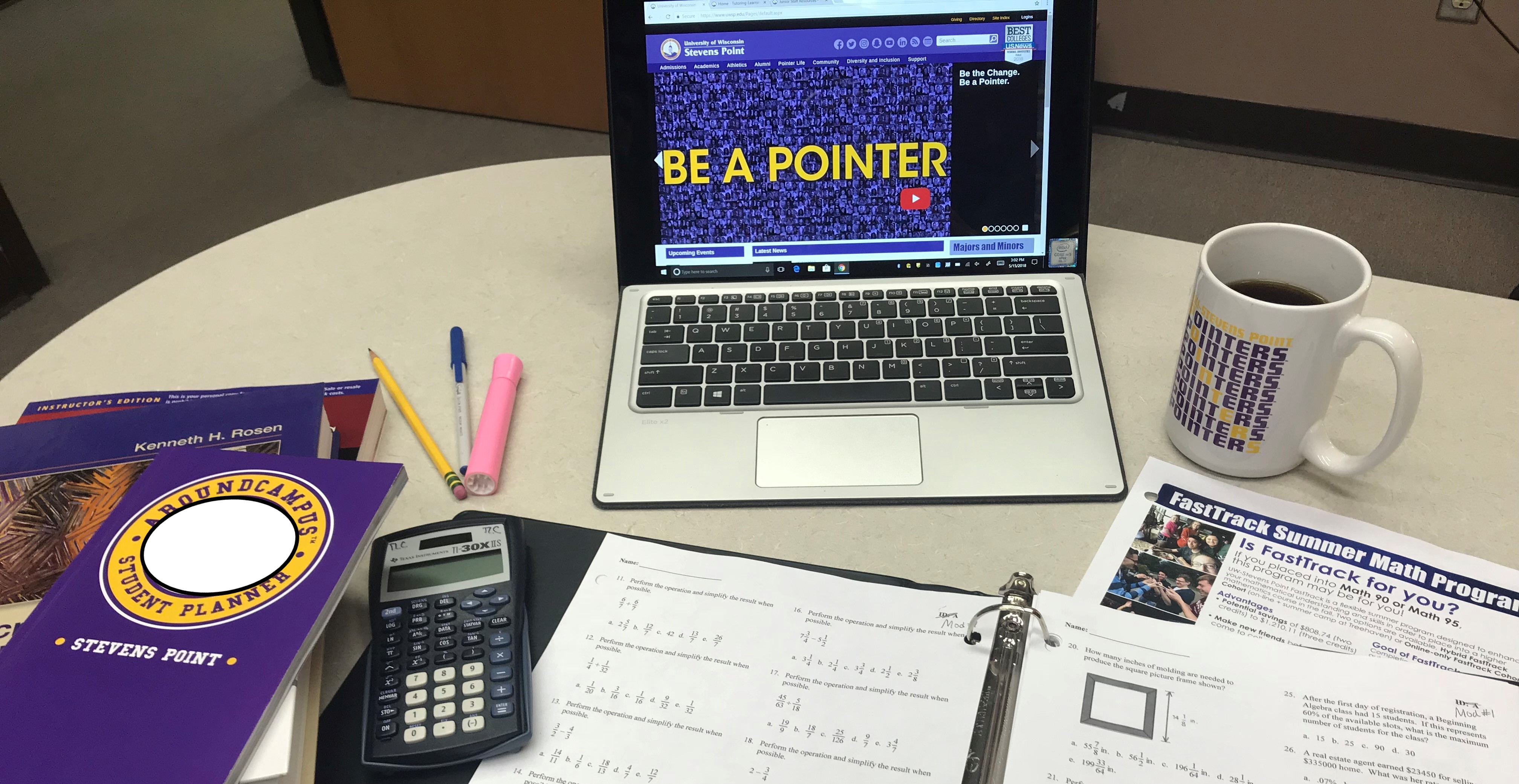 Various items are spread on a tabletop: a purple Stevens Point planner on top of textbooks, a pencil, a blue pen, a pink highlighter, a calculator, a binder opened to pages with math problems on them, a laptop showing the UWSP homepage, and a Pointers mug with coffee in it.
